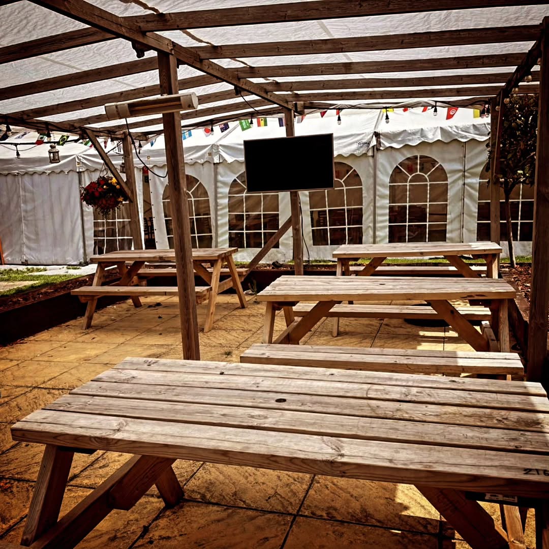 Wooden picnic benches under a covered pergola with overhead heaters, bunting and festoon lights, looking towards a white marquee with arched windows