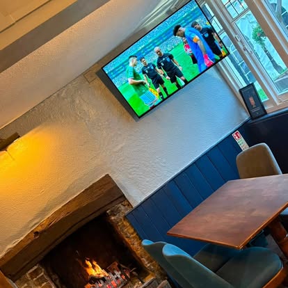 A lit open fire in a brick fireplace beside a wooden table and upholstered chairs, with football showing on a wall-mounted screen above